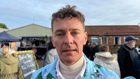 Tom wears a Christmas shirt over a polo neck. Shoppers can been seen behind him. He has short, curly brown hair and is clean shaven.