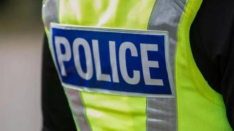 A police officer (photographed from behind) wearing a yellow high visibility jacket which has 'Police' written on it in white lettering on a blue background. 