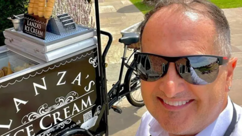 A man with short dark hair and mirrored sunglasses with a black box next to him with Lanza's ice cream on the side