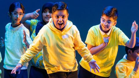 Boy in yellow top very expressive looking to camera surrounded by boys in yellow and girls in pale green

