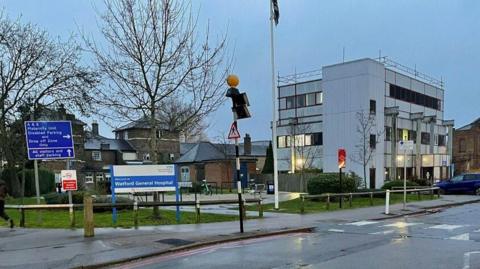 An entrance to Watford General Hospital, showing a Watford General Hospital sign in white lettering on a blue background. Another sign gives directions for various car parks. There is a three-storey white building to the right, with black window frames and some windows lit up. There is a two-storey brick building in the background, and a road runs past in the foreground.