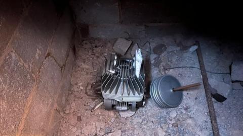 A small motor engine on a rough concrete and gravel floor inside a dimly lit brick enclosure under the floorboards of a house in Devon.