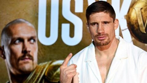 Rico Verhoeven holds up one finger and stands in front of a board with an image of Oleksandr Usyk