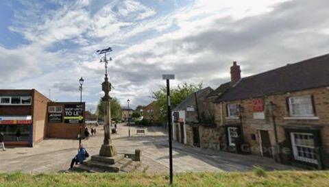 The pedestrianised Market Square in Woodhouse, Sheffield with an old market stone cross/war memorial in the centre. A man in blue with a rucksack is sitting on the steps of the cross. An old stone building, a pub, is to the right of the picture with shops to the left. A hedge is in the foreground of the picture.