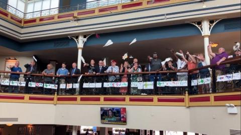 A group of people are standing on a balcony and can be seen launching paper aeroplanes into the air.