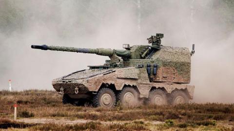 A large military vehicle with four wheels on one side. It is covered in brown and green camo paint. On top, there is a long thin gun barrel. There is smoke all around it