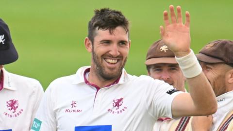 Craig Overton celebrates taking a wicket. He is smiling and holding his hand up.