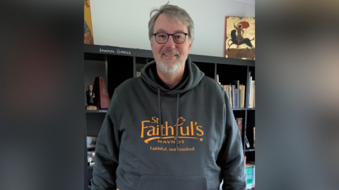 A man wearing a hoodie saying 'St Faithful's Havnot. Faithful, not finished'. He is smiling at the camera and has grey hair and a beard and wears black framed glasses.