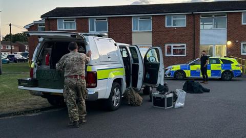 A soldier standing behind a military vehicle, which is parked facing the front of a row of terraced houses. A police car is parked at a right angle in front of  one of the houses and a man dressed in black is standing by the passenger door