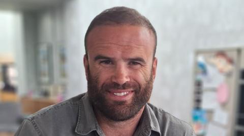Former rugby player Jamie Roberts, with short cropped brown hair and a beard, wearing a grey shirt, smiles at the camera