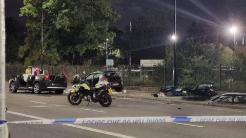 Car behind police tape with roof cut off. Police bike and officer can be seen along with debris