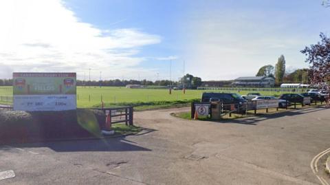 A view of the entrance to the Cambridge Rugby Football Club. A large sports field can be seen just beyond a small car park, in which cars are parked. 