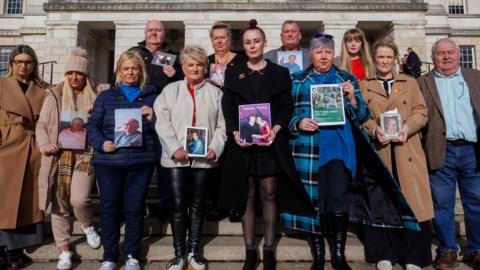 Members of the Northern Ireland Covid Bereaved Families for Justice group on the steps of Parliament Buildings at Stormont.

They are holding pictures of their family members and looking directly at the camera. 
The back row left to right:
male (bald in a black coat), female (blond, black top), male (grey short hair and grey suit jacket), female (blond, long hair and red top)
The bottom row left to right:
female (blond middle parting, glasses and brown coat), female (beige hat and coat with scarf and blond long hair), female (blond shoulder length hair and navy coat), female (cream zip up and short blond hair), female (black coat and red hair tied up), female (blue hair short and blue coat and scarf), female (brown coat and blond hair) and male (grey hair with blue shirt and tweed brown jacket). 

