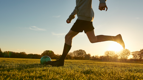 Generic image of a footballer from the waist down dribbing a ball on a field. they are wearing dark boots and socks and dark shorts, with a light grey top
