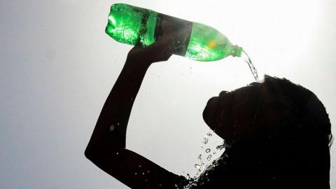 A young woman with long hair is silhouetted against the sun as she pours water on her face from a green plastic bottle held above her head. 