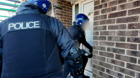 Police in riot gear bash down a white door
