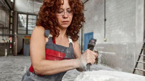 A person wearing glasses and a protective apron stands in a large indoor workshop, using a hammer and chisel to shape a block of pale stone. Fine dust is visible and the surface of the stone is rough and partially formed. The background shows a high‑ceilinged industrial space with white walls, exposed fixtures, and a ladder leaning against the wall.