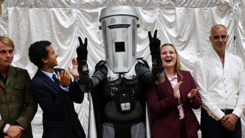 Man wearing grey bin mask over his head lifting two arms and holding his fingers in peace signs