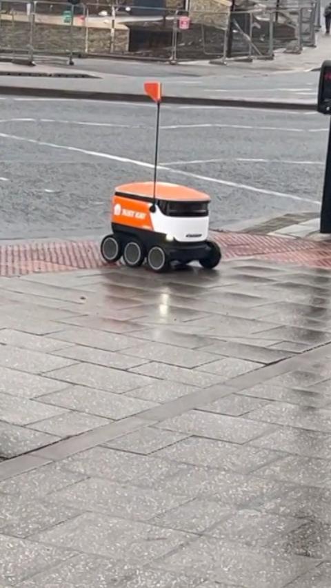 A Just Eat-branded orange and white delivery robot crossing a pedestrian crossing. It has six wheels and a pole reaching out from the top with an orange flag on the top. 