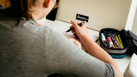 An unidentifiable young person with brown hair is wearing a grey top while sitting at a desk and writing on a squared notepad. They have a pencil case filled with stationery items.