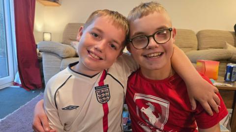 Two young boys with blonde hair are wearing England and Arsenal football t-shirts standing arm in arm. They are in a living room and both smiling at the camera. Josh, on the left, is wearing dark-rimmed glasses. Behind them is a beige-coloured sofa.