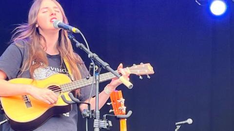 Amelia Coburn plays a guitar and is singing on stage. She wears a dark-coloured T-shirt.