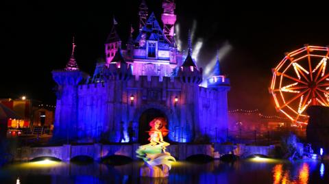 A wide shot of Banksy's Dismaland at night, showing a Disney-like castle lit in purple and a Little Mermaid-like statue