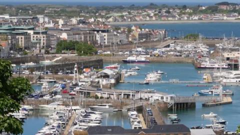 A large marina with assorted boats and buildings behind along with a beach and more houses.