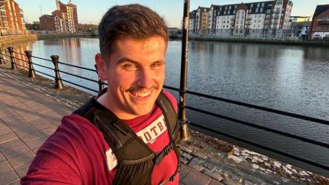 A man with short brown hair wearing a red tshirt and a black running vest, he is smiling at camera as he takes a selfie, running alongside the water in Belfast. There are high rise waterfront apartments visible on the horizon.