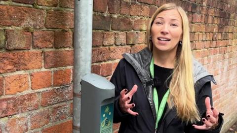 A woman with long blonde hair and wearing a black waterproof coat and a green lanyard, standing next to a lamppost with a grey boxed attached to it. There's a brick wall behind her. She appears to have been photographed mid-sentence and is gesturing with her hands.