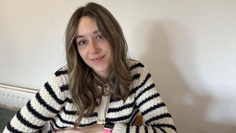 Abbi, a young woman with dark blonde hair. She is wearing a black and white striped cardigan and is sitting at her kitchen table