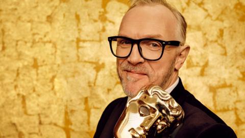 Greg Davies is pictured holding a Bafta TV award. He is wearing glasses and a black suit jacket and white shirt. The background is gold.