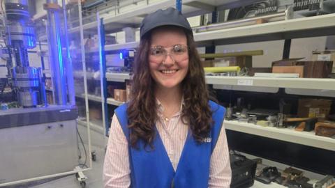 A woman wearing a blue vest, safety goggles and a black hat. She is standing in front of shelving containing various bits of equipment, and is smiling
