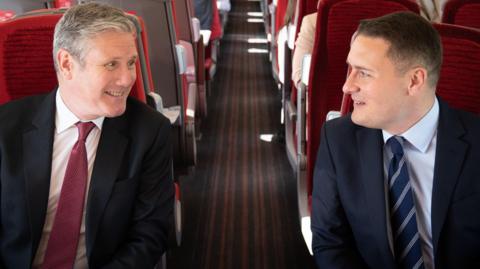 The PM sits across a train aisle from Wes Streeting, both smiling and looking at each other. The PM's tie is red, matching the seats and Streeting's is navy blue.