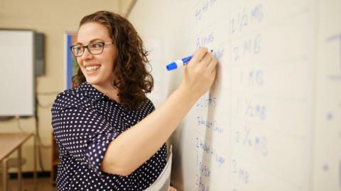 Teacher with shoulder-length brown curly hair and wearing glasses stands with a blue marker writing on a whiteboard. Stock image.