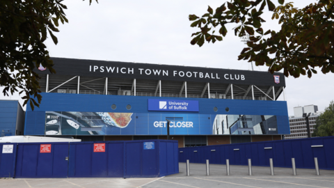 Ipswich Town Football Club's Portman Road stadium with the University of Suffolk sponsorship logo on the side of the stand. Trees surround the picture