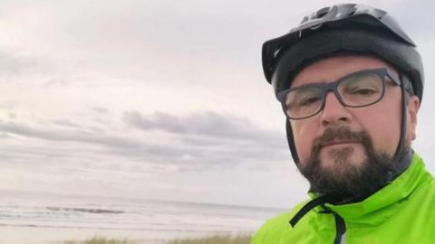 Ian Price, who has a beard, is wearing glasses and a cycle helmet, for a selfie at a beach, in which he is also wearing a hi-vis jacket