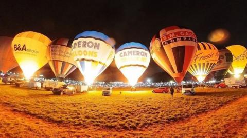 Nine hot air balloons in a row illuminated against the night sky  with vehicles parked on a grassy field in the foreground and the night lights of Bristol in the background