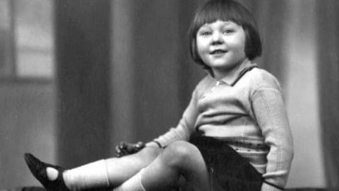 Black and white image of Ruby Currell as a five-year-old sitting and smiling at the camera