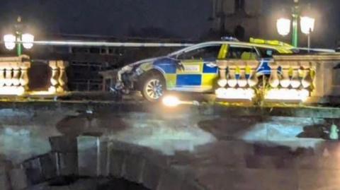 A police car that has driven through a balustrade on a bridge. The car is partially overhanging the bridge edge. It is night and the scene is lit by lampposts.