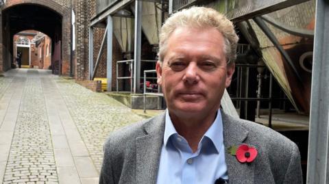 Shepherd Neame chief executive, Jonathan Neame is wearing a grey jacket and light blue shirt and is standing in front of the brewery