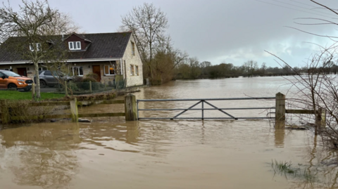 A field covered in floodwaters with a partially submerged gate and a home in the background which has remained just above the water