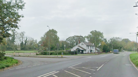 A junction on the A134 on Lynn Road. In the distance past the turning is a white property and a blue van driving away.