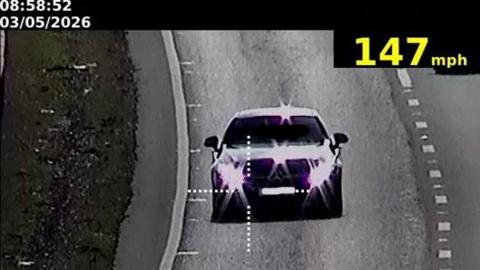A black and white CCTV-style image of a Mercedes with the light reflecting off its lights and bonnet driving on the A14.