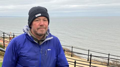 Phil Gallon is standing on a beach promenade and looking into the camera. He is wearing a grey woolly hat and a blue jacket.