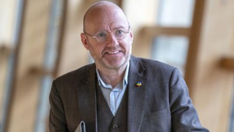 A smiling bald man with glasses, wearing a grey three-piece suit and blue shirt, looks slightly to his left while walking in the Scottish Parliament 