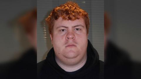 Police custody photograph of David Thorne who has red wavy hair and is wearing a dark coloured top