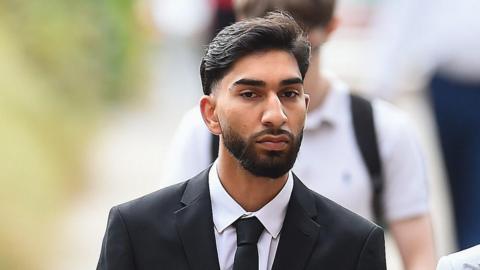 A young man with a dark beard wears a white shirt and a black tie and jacket and walks along a street. 