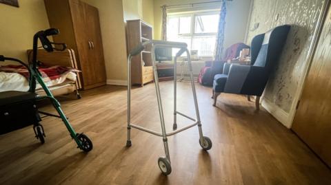A room in a care home, featuring a bed, chairs, zimmer frame and other furniture. 