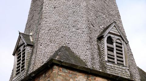 A close up shot of the lower part of a church spire, back by a blue sky. There are small oak panels covering the spire, with wooden, worn through windows.